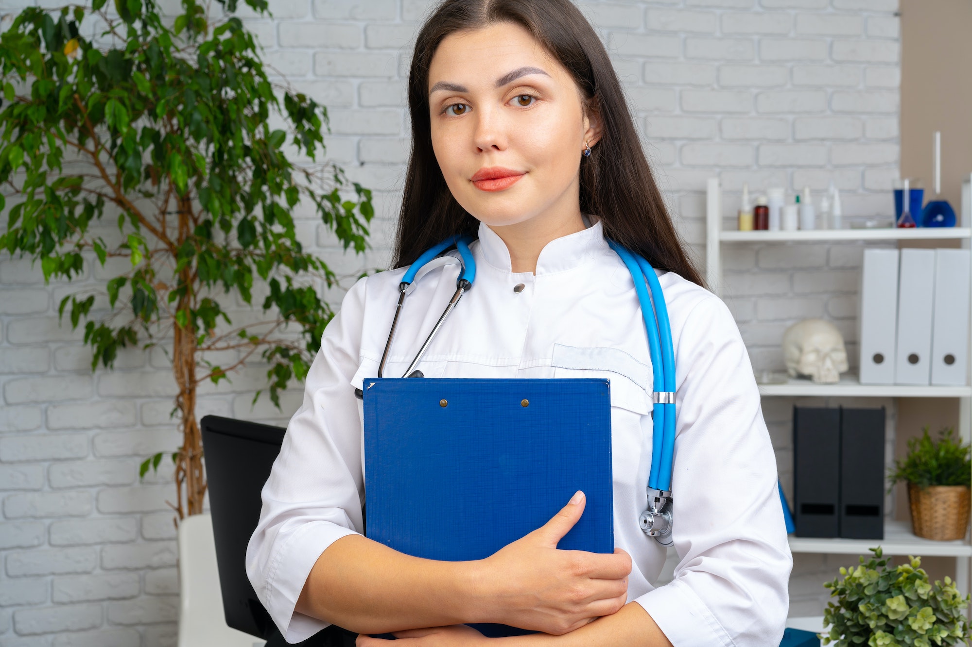 Young brunette woman doctor standing with clipboard in her office